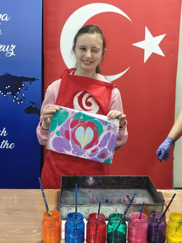 elevă zâmbește și arată lucrarea realizată - o inimiă colorată acvamarin și roz