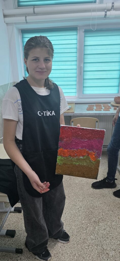 Elevă arată planșa realizată, cu dungi orizontale groase colorate roșu, roz, oranj, verde, maro deschis.