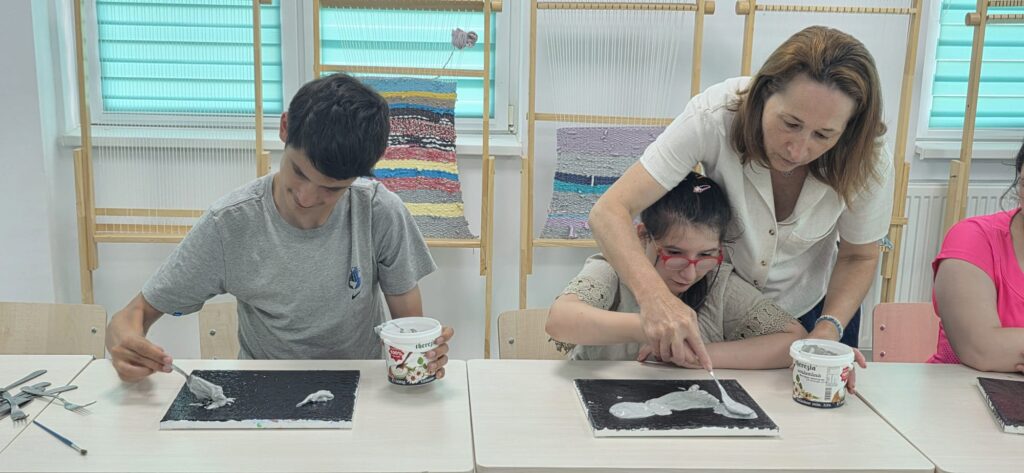 Doi elevi și doamna profesoară întind ciment lichid pe planșele lor folosind linguri