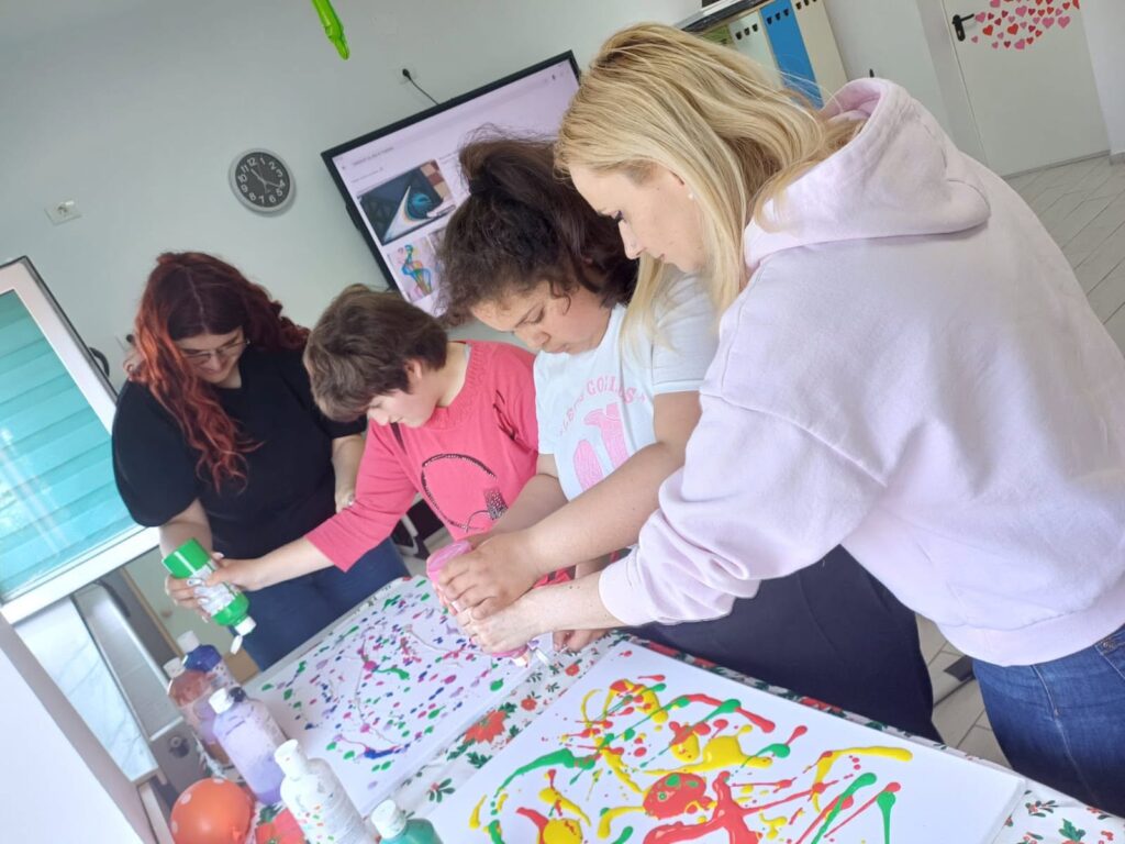 Doamna profesoară sprijină elevii, în fața planșelor mari albe cu dungi și puncte de vopsea colorată