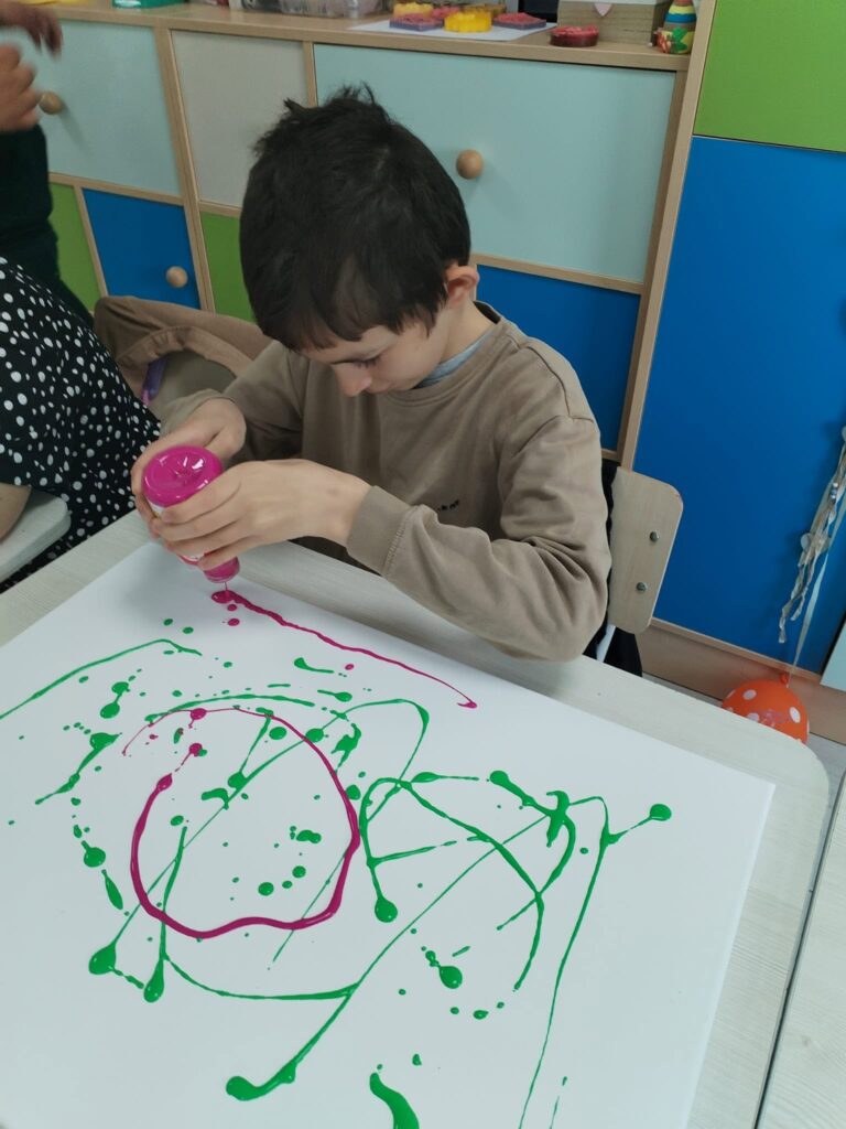 Elev ține balonul roz și pictează linii colorate pe o planșă mare albă