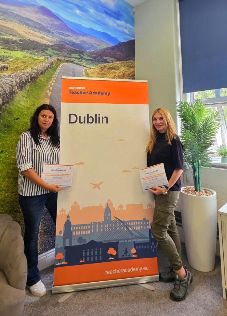 Cele două profesoare țin certificatele de absolvire, de o parte și de alta a bannerului „Europass Teacher Academy – Dublin”.