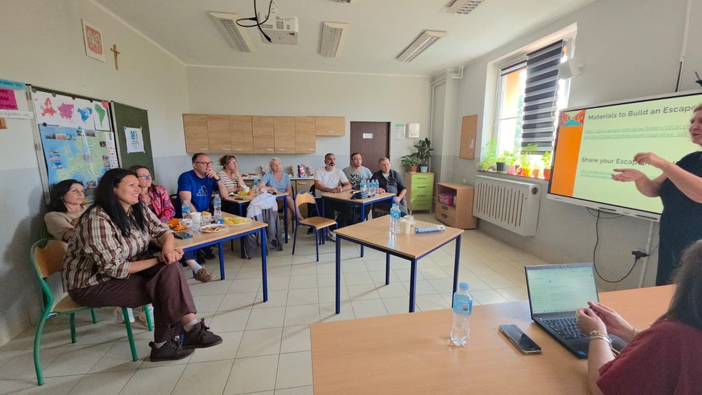 Cadrele didactice stau la mese și urmăresc o prezentare despre materialele necesare pentru construirea unui escape room educațional. Formatoarea explică ce apare pe tabla interactivă.