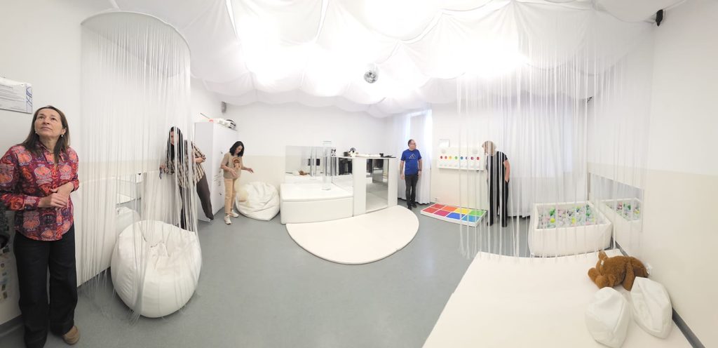Imagine panoramică a camerei senzoriale, decorată în alb: fotolii-sac, perdele din fire, un podium cu oglinzi, un panou de perete cu butoane colorate, un bazin cu bile și o saltea cu un ursuleț de pluș. Mai multe cadre didactice privesc echipamentele.
