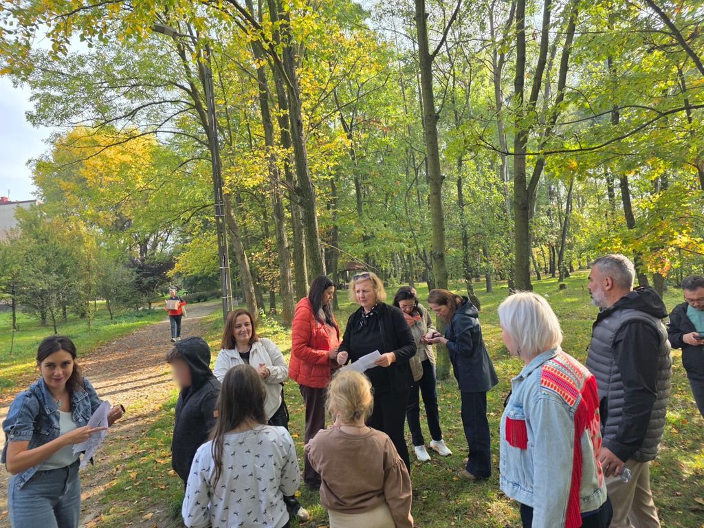 Cadre didactice și elevi adunați într-un parc cu copaci înalți, în culori de toamnă. O profesoară explică sarcinile, iar participanții citesc fișe de lucru.