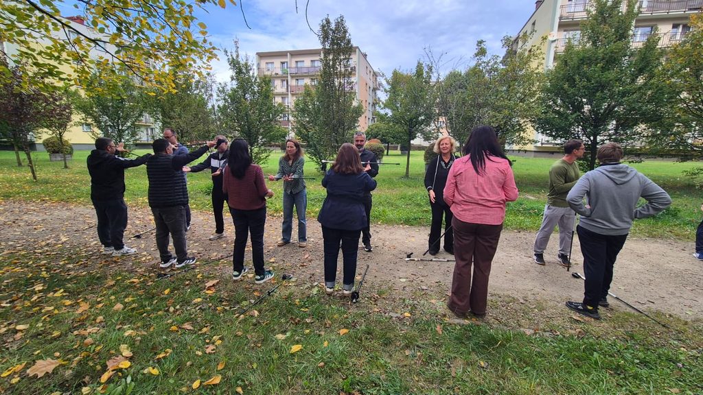 Cadrele didactice pe o alee din parc, între blocuri, și exersează în perechi ținând bastoanele de Nordic Walking orizontal, întinse între ei. O instructoare explică mișcările.