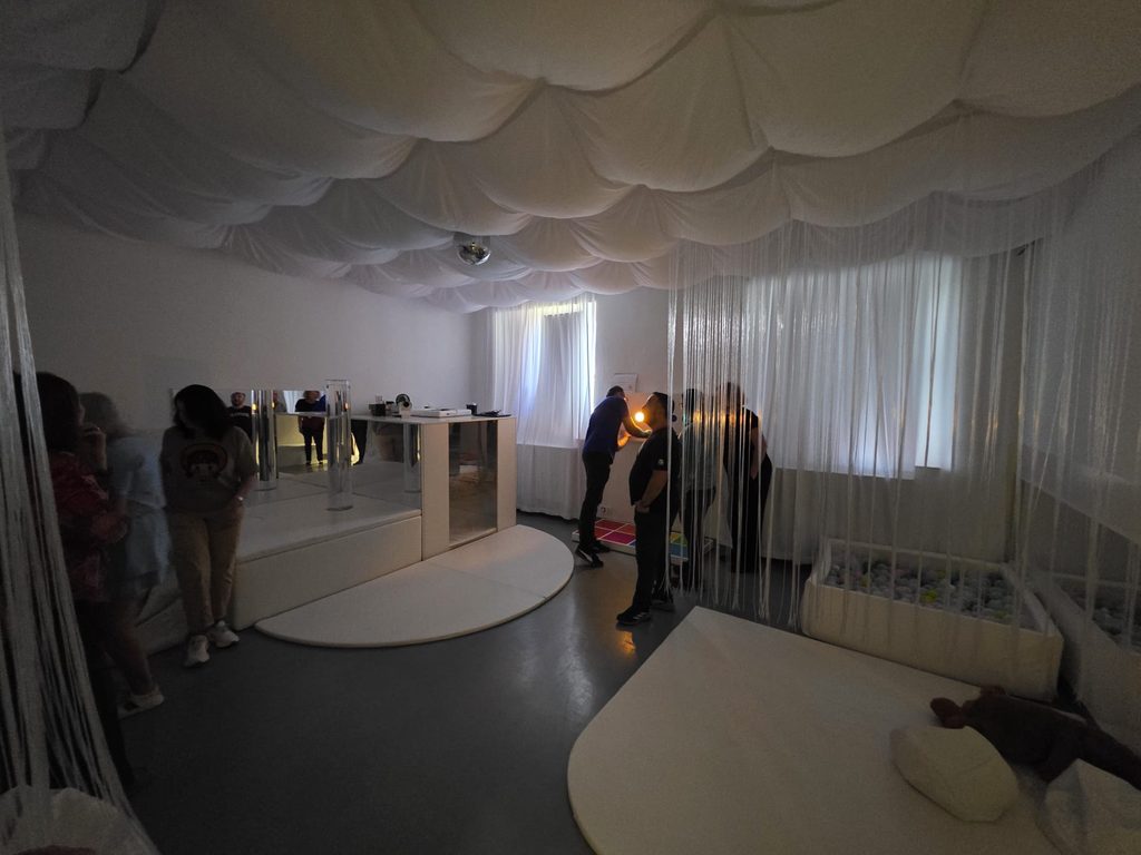 Participanții explorează camera senzorială cu luminile stinse. Tavanul este acoperit cu draperii albe, iar în centru atârnă o sferă cu oglinzi. Lângă un panou interactiv de pe perete, un profesor ține o mică sursă de lumină caldă. În dreapta se văd perdeaua din fire și bazinul cu bile.