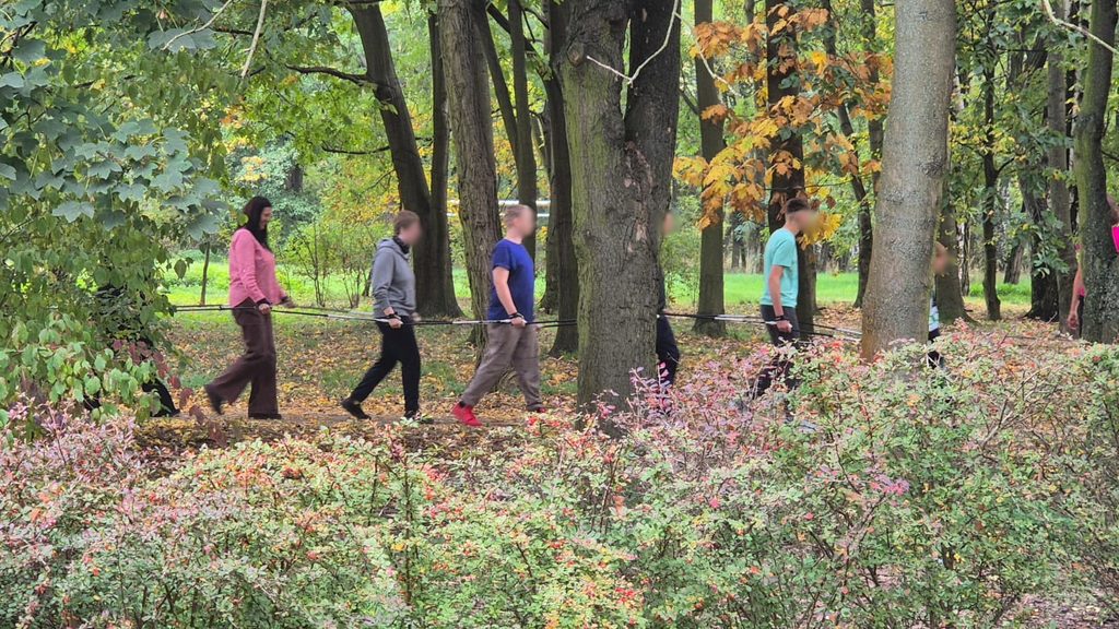 O profesoară și mai mulți elevi merg în șir printr-un parc cu copaci tomnatici. Fiecare ține de bastoanele celui din față, formând un lanț. În prim-plan sunt tufe cu frunze roșiatice.