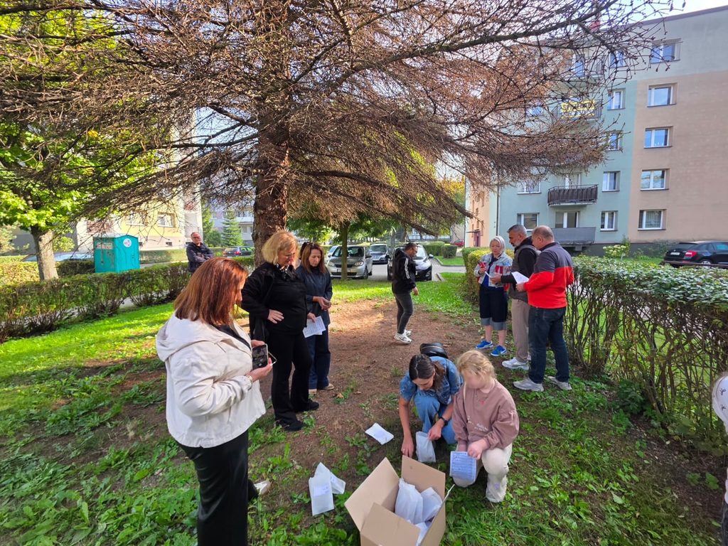 Profesori și elevi lângă un copac, între blocuri. O profesoară și o elevă, aplecate lângă o cutie, folosesc pungi de hârtie și fișe pentru a strânge materiale naturale.