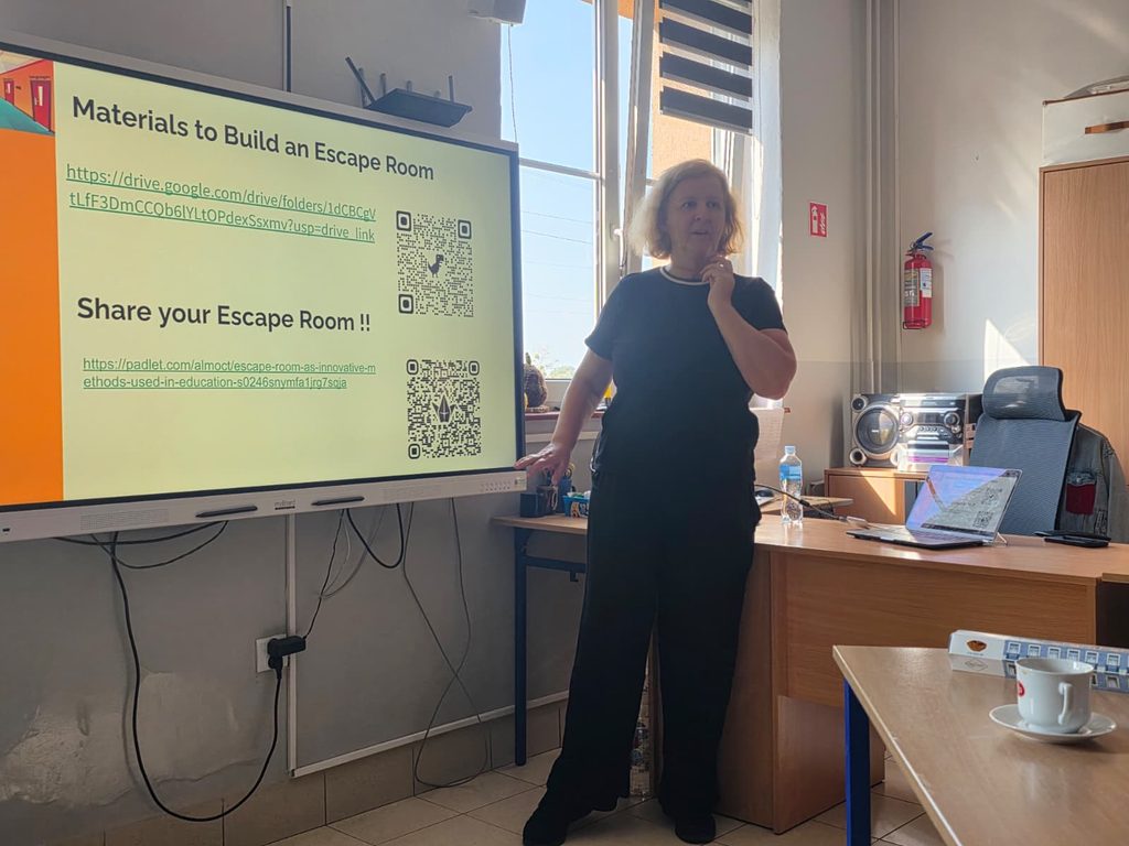 Formatoarea stă lângă tabla interactivă, pe care este un slide cu titlurile „Materials to Build an Escape Room” și „Share your Escape Room!!”. Slide-ul conține linkuri și coduri QR către un dosar cu materiale și către un panou Padlet, unde participanții își pot publica jocurile.