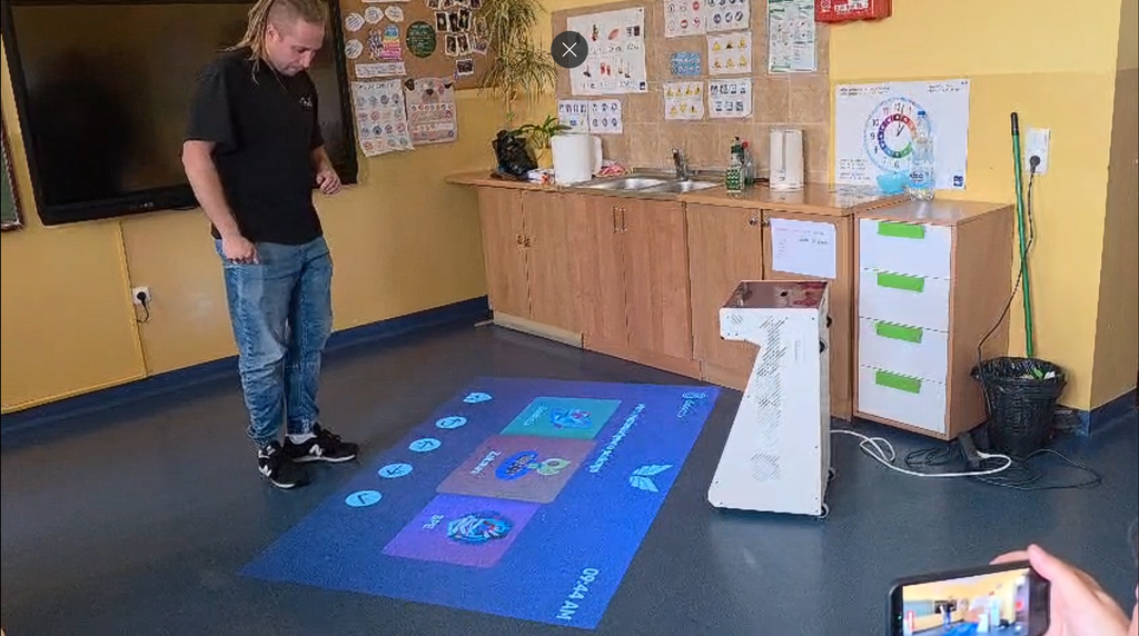 Un profesor stă lângă o proiecție interactivă pe podea, care afișează un meniu de jocuri educative. Lângă el se află aparatul de proiecție, iar pe pereți sunt materiale didactice.