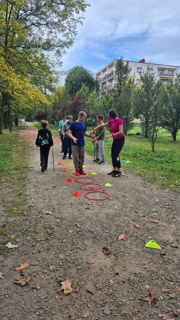 Elevi cu bastoane de Nordic Walking parcurg un traseu cu cercuri colorate și conuri, pe o alee din parc. O profesoară ghidează un elev printre obstacole.