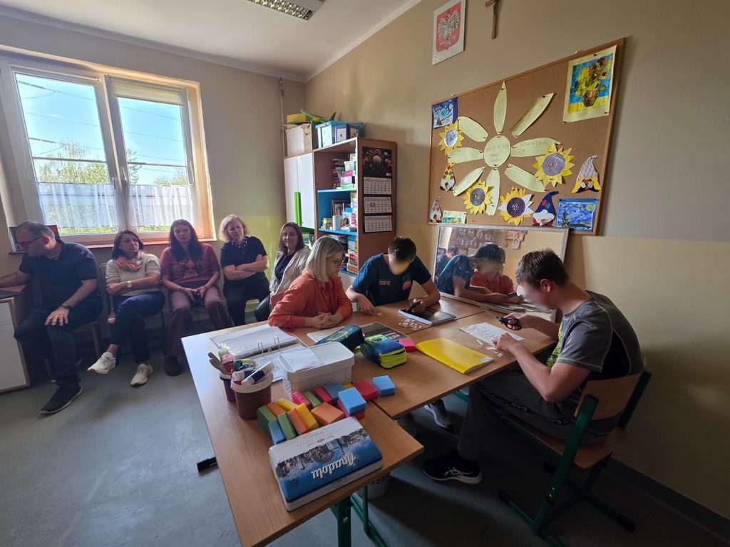 Doi elevi lucrează la o masă cu o profesoară logoped, decupând și potrivind cartonașe. Pe masă sunt bureți colorați și materiale de lucru. Cadrele didactice din proiect observă lecția de pe scaune, lângă fereastră.