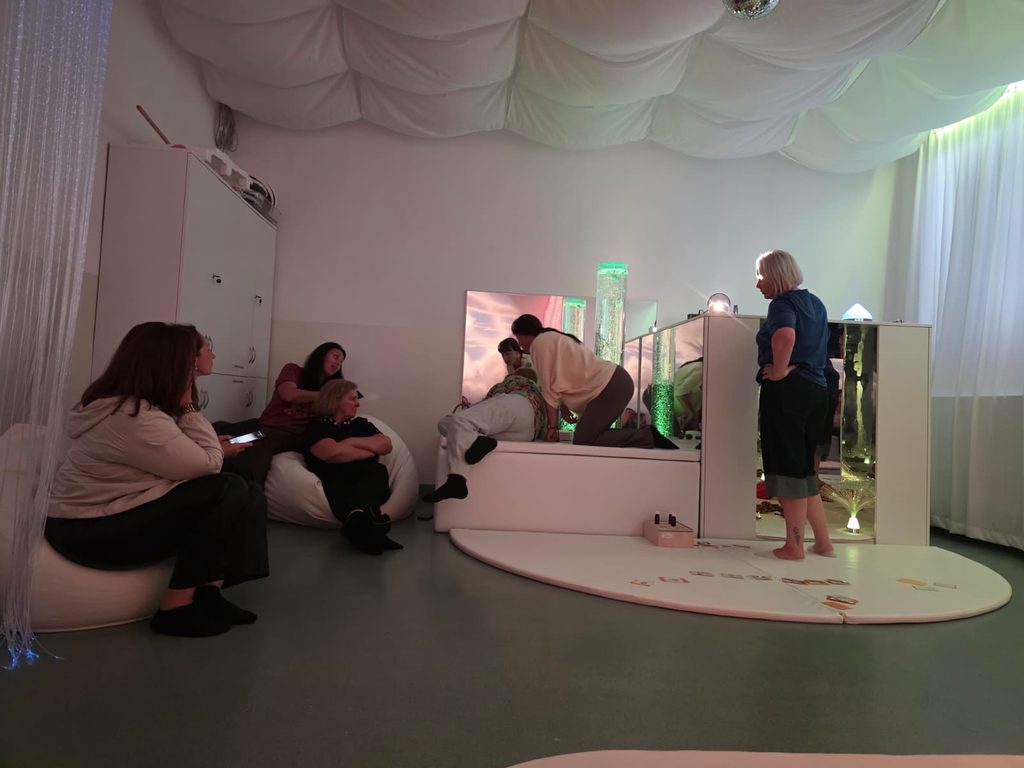 Lecție în camera de stimulare senzorială - Snoezelen, în lumină difuză, verde, de la o coloană cu bule și de la fibrele optice. Pe podium, o profesoară lucrează cu un elev. Cadrele didactice observă activitatea, așezate pe fotolii-sac.