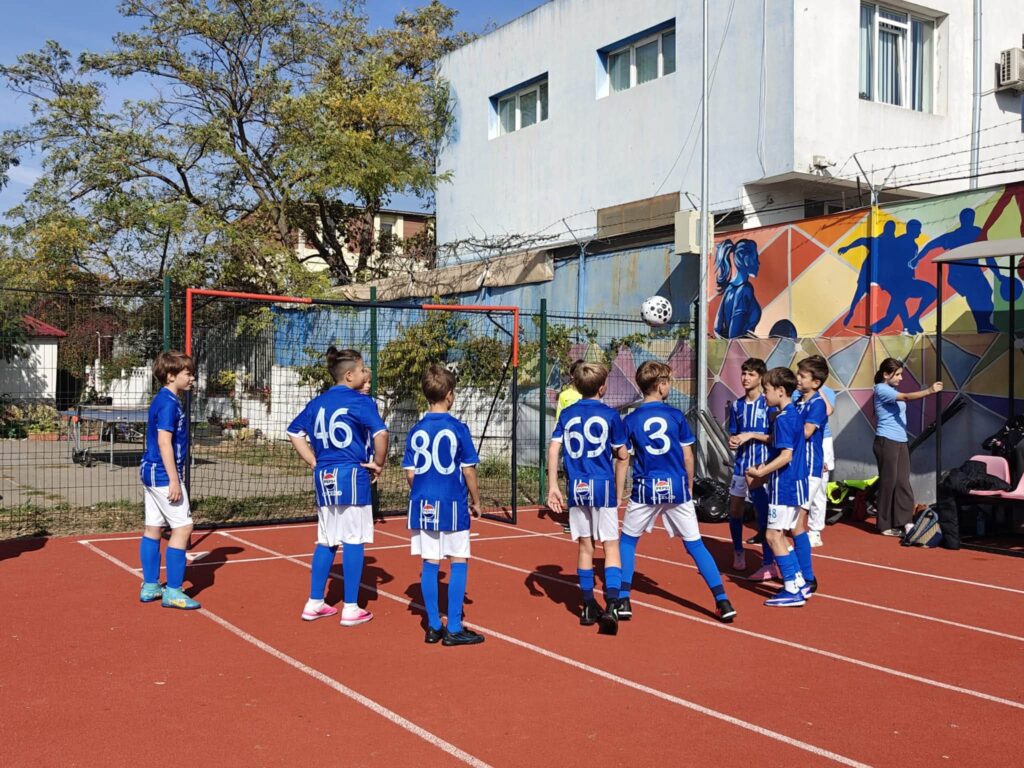 Tineri fotbaliști în echipament albastru se încălzesc cu mingea înaintea unui meci amical pe terenul școlii.