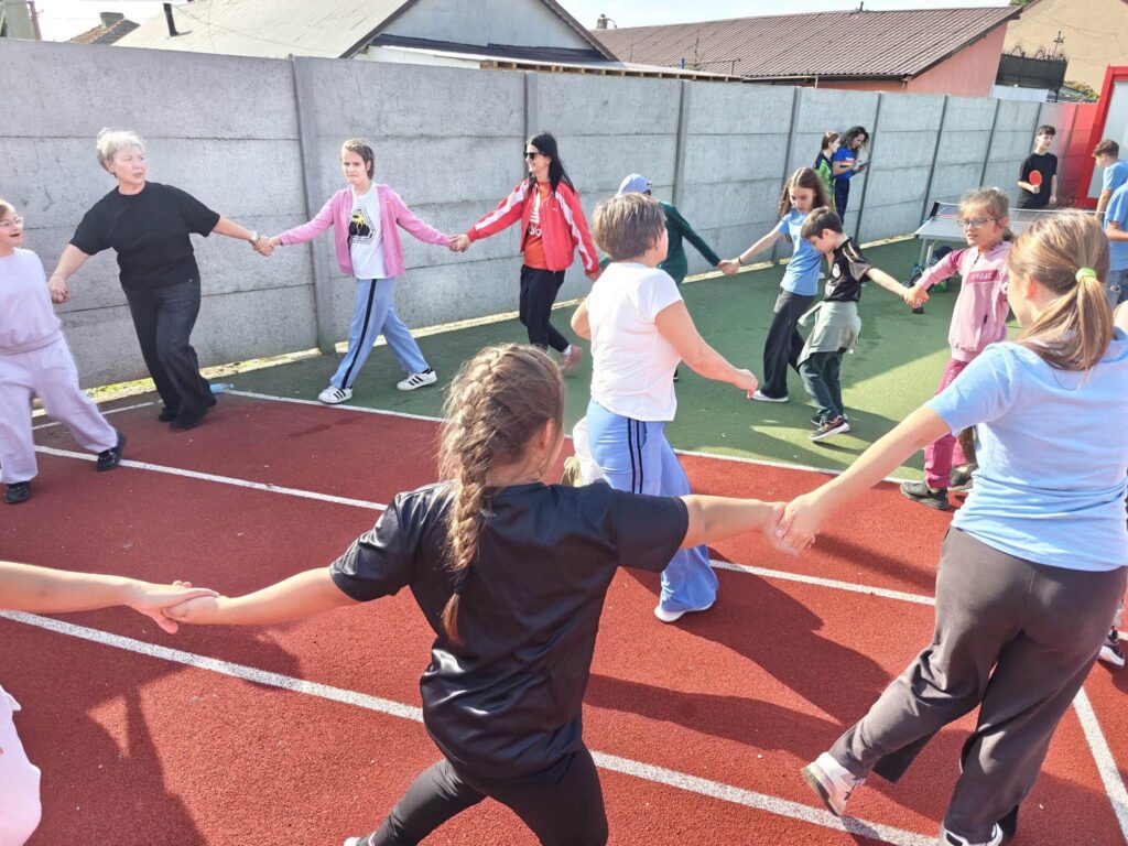Elevi și cadre didactice se țin de mână într-o horă mare; în fundal, un elev joacă tenis de masă.