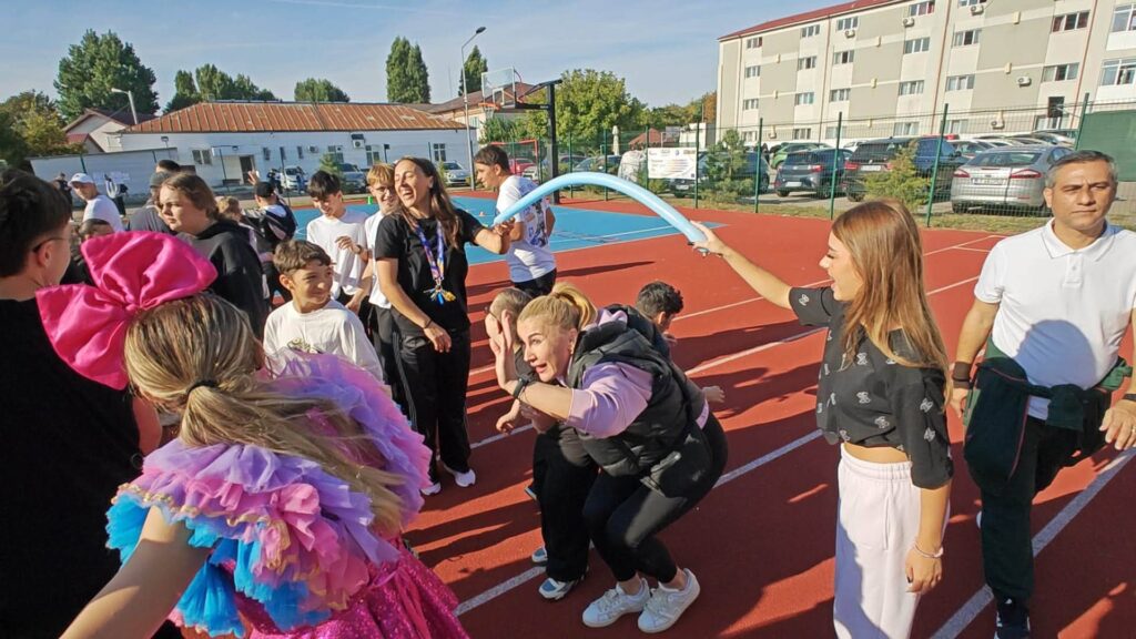 O profesoară trece pe sub un balon lung albastru într-un joc de tip limbo, încurajată de elevii din jur.