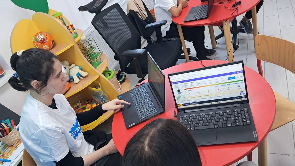 O elevă arată detaliile de pe ecranul unui laptop în timp ce colaborează cu un coleg la o masă de lucru.